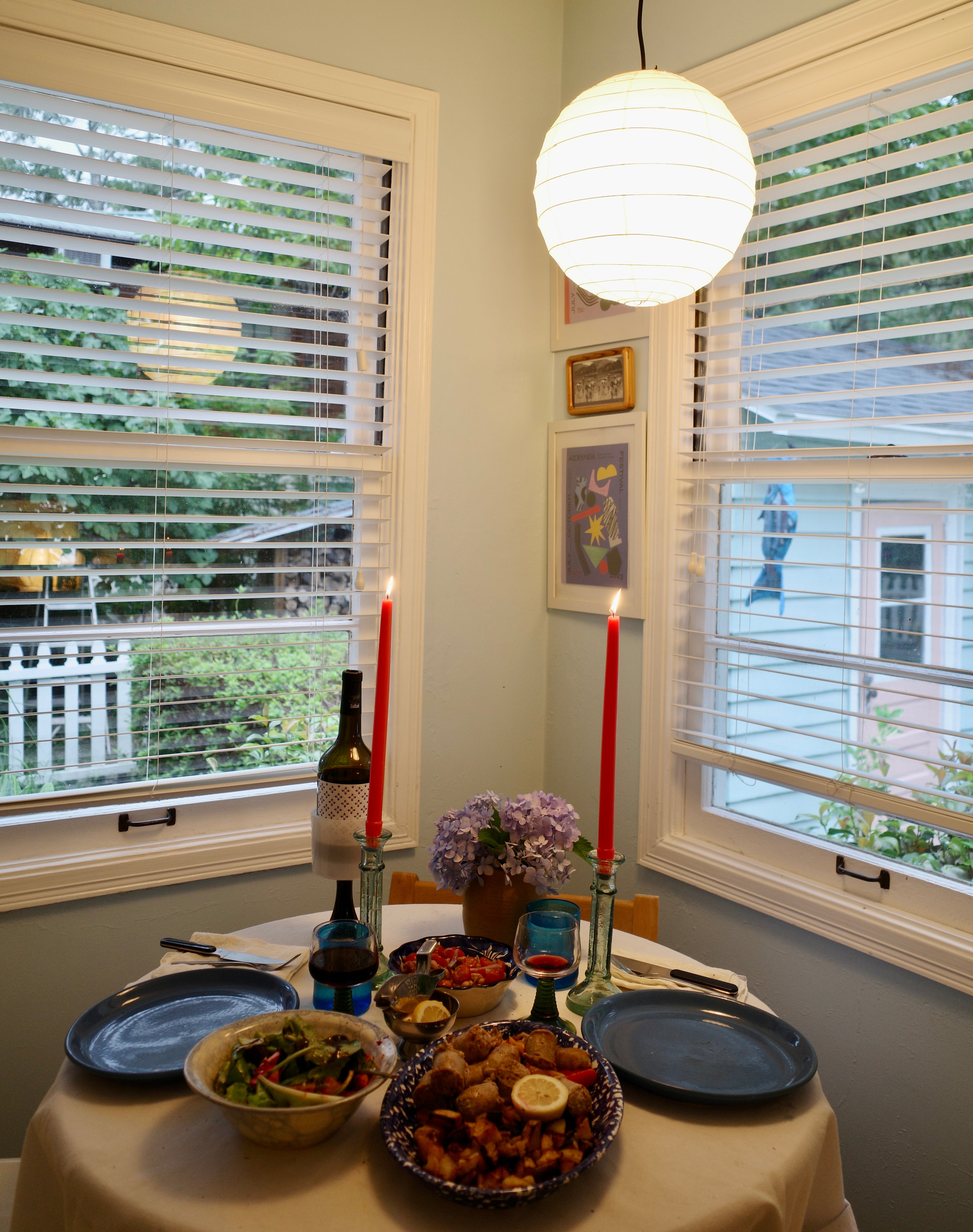 Dining table set for a meal with plates, bowls, and candles in a room with windows.